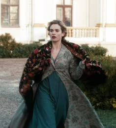 a woman in a long dress and coat walking down the street with her hand on her hip