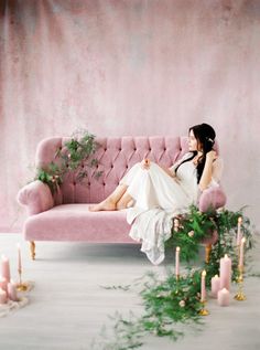 a woman sitting on top of a pink couch next to candles and greenery in front of a wall