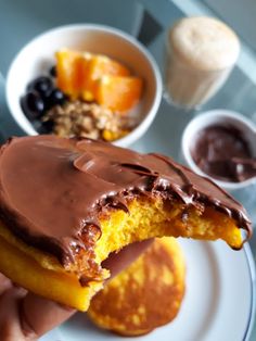 a person holding up a piece of cake with chocolate frosting and fruit in the background