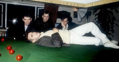 three men are playing pool in a room with balls on the floor and one man is laying down