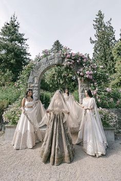 three women dressed in wedding gowns standing next to each other near an arch with flowers on it