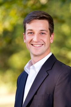 a man in a suit and white shirt smiling at the camera with trees in the background