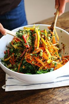 a person is holding a wooden spoon over a bowl of food that includes carrots, lettuce and other vegetables