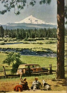 two people sitting on the ground next to an old truck and trees with a mountain in the background