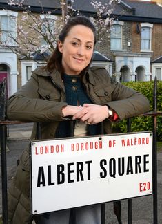 a woman standing next to a sign that says albert square on the street in front of some houses