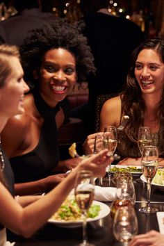 three women sitting at a table with wine glasses in front of them and plates of food on the table