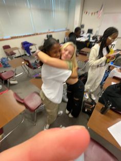 two girls hugging each other in a classroom with desks and people standing around the room