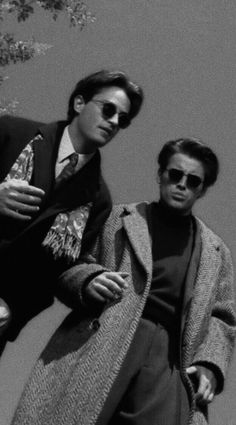 two men standing next to each other in front of a tree and one is wearing sunglasses