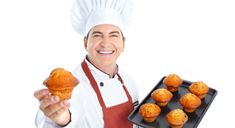 a man in an apron and chef's hat holding a tray with muffins on it