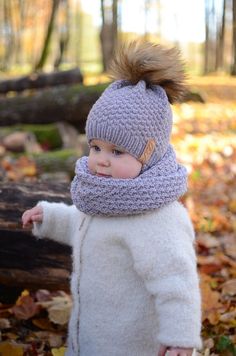 a small child wearing a knitted hat and scarf in the woods with leaves on the ground