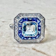 Breathtaking handcrafted platinum Art Deco style aquamarine, sapphire and diamond ring! The center stone is a lovely 1.05 carat aquamarine. It is surrounded by a row of fine French cut sapphires weighing 0.70 carat. The sapphires are haloed by 0.40 carat of round diamonds. Fine milgrain adds to the delicacy of the ring. Beautiful openwork filigree decorate the under gallery. The ring measures 13.97 x 13.63 mm from side to side. Stamped Pt900. Ring size 6.25-6.5 US / M-M 1/2 UK. > Resizing includ Blue Platinum Art Deco Diamond Ring, Blue Emerald Cut Diamond Ring With Diamond Accents, Blue Emerald Cut Diamond Ring With Accents, Blue Diamond Ring With Diamond Accents Emerald Cut, Blue Diamond Art Deco Ring, Blue Art Deco Diamond Ring, Blue Art Deco Jewelry Gia Certified, Art Deco Blue Diamond Ring, Blue Art Deco Diamond Ring With Center Stone