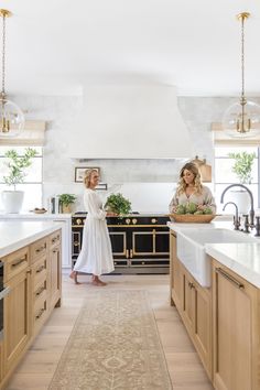 two women standing in a large kitchen with white counter tops and wooden cabinets, along with an area rug on the floor