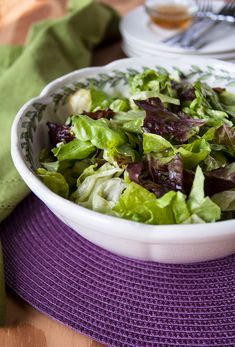 a white bowl filled with lettuce on top of a purple table cloth next to silverware