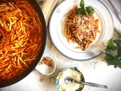 two plates with pasta and sauce on them next to a bowl full of basil leaves