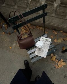 a person is sitting on a bench reading a newspaper and holding a brown handbag