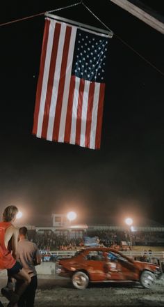 an american flag hanging from the side of a building next to a red car at night