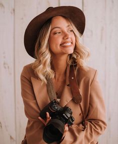 a woman wearing a brown hat and holding a camera in her right hand while smiling at the camera