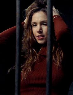 a woman in a red sweater behind bars looking at the camera with her hand on her head