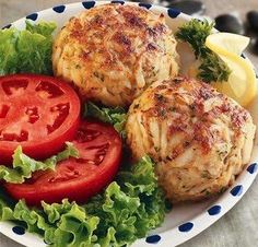 two crab cakes with tomatoes and lettuce on a plate next to lemon wedges