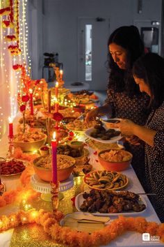 two women standing in front of a long table filled with plates of food and candles