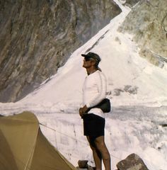 a man standing next to a tent on top of a snow covered mountain in the mountains