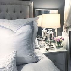 a white bed topped with pillows next to a night stand and two vases filled with flowers