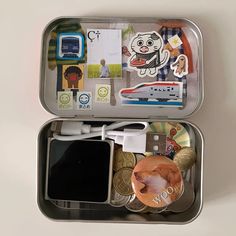 an open tin with various items in it on a white surface, including a cell phone and some coins