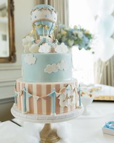 a three tiered cake is decorated with blue and pink stripes, clouds, and a hot air balloon