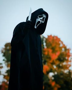 a person wearing a mask and holding a knife in front of trees with orange leaves