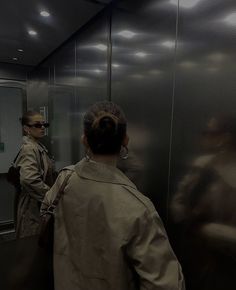 two women standing in front of an elevator, one is wearing a trench coat and the other has sunglasses on her head