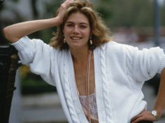 a woman leaning against a pole wearing a white sweater and pearl necklace, with her hands on her head