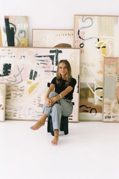 a woman sitting on top of a chair in front of some paintings and artwork with her legs crossed