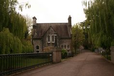 an old stone house with trees lining the street and fenced in area next to it