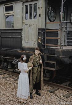 a man and woman standing next to a train car on the tracks with an old fashioned locomotive in the background