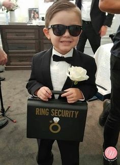 a little boy dressed in a tuxedo and holding a ring security box with a flower