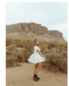 a woman in white dress and black boots standing on dirt road with mountains in the background