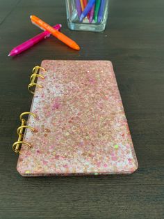 a pink notebook sitting on top of a wooden table next to markers and pencils