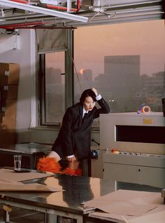 a woman in a black suit and orange skirt leaning on a table next to a window