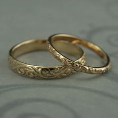 two gold wedding rings sitting on top of a gray surface with swirly designs in the middle