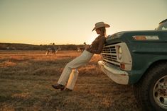 a woman leaning on the hood of a truck in a field with horses behind her