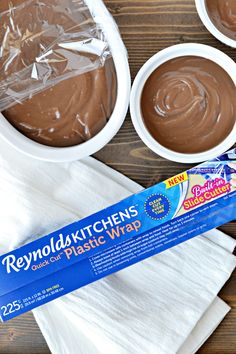 three bowls of chocolate frosting on top of a wooden table with plastic wrap around them