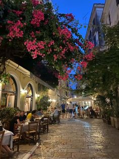 people sitting at tables in an alleyway with pink flowers on the trees above them