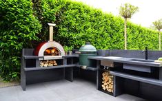 an outdoor bbq grill with firewood in the middle and green bushes behind it