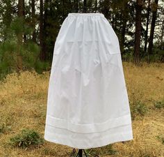 a woman's white dress is standing on a tripod in the grass with trees behind her