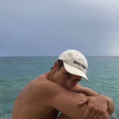 a shirtless man sitting on the beach with his arm around his chest and looking at the ocean