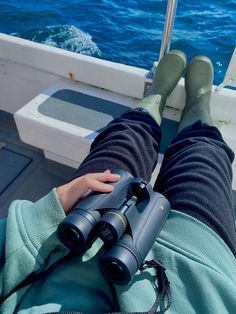 someone is sitting on a boat looking at the water with their legs crossed and binoculars in hand