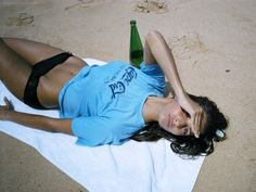 a woman laying on top of a towel next to a bottle of beer in the sand