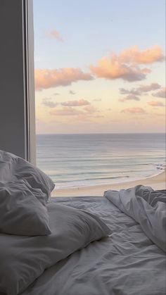 an unmade bed sitting in front of a window overlooking the beach and ocean at sunset