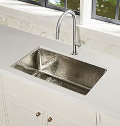 a stainless steel sink in a white kitchen with marble countertops and windows behind it