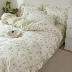 an unmade bed with white sheets and flowers on it, next to a potted plant